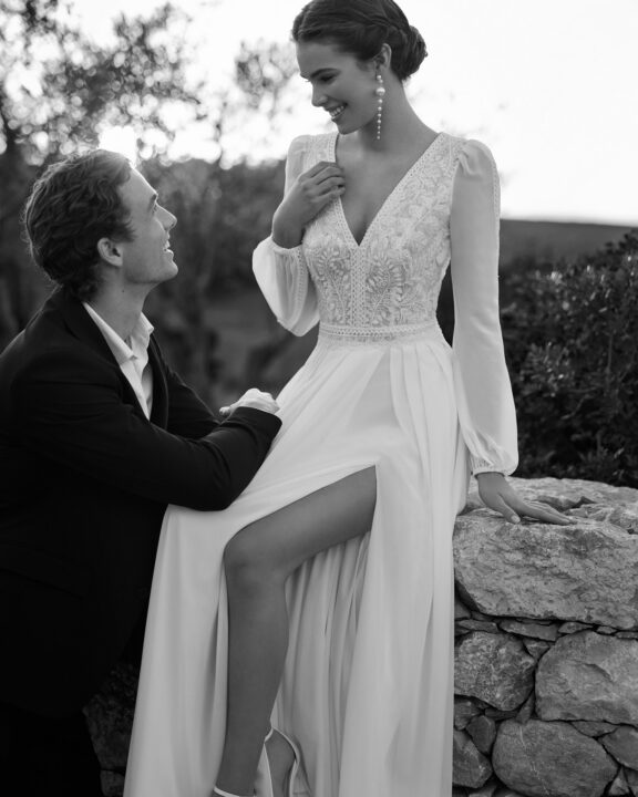 Elegante trouwjurk van Rosa Clará met lange mouwen, diepe V-halslijn, romantische kantdetails en een hoge split. Deze jurk heeft een prachtige open rug met delicate kant en een vloeiende rok met een lange sleep, perfect voor een moderne en verfijnde bruid in België.