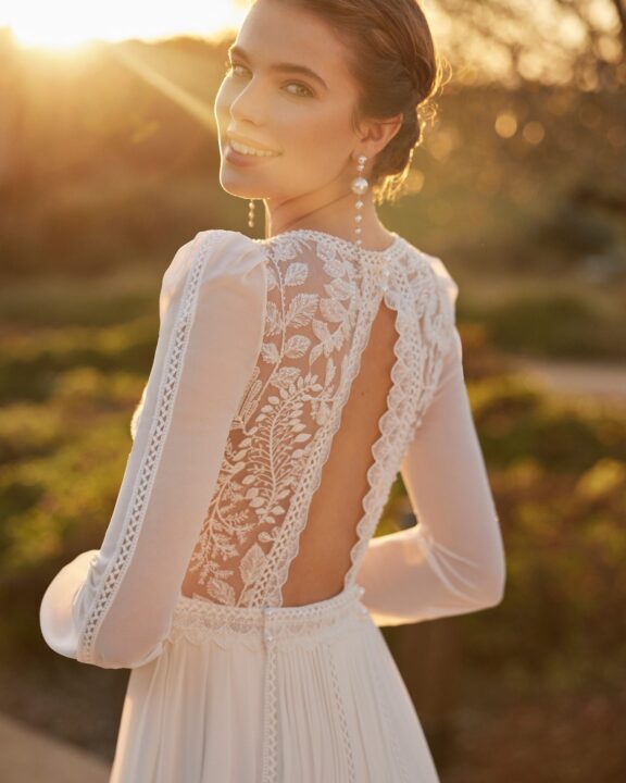 Elegante trouwjurk van Rosa Clará met lange mouwen, diepe V-halslijn, romantische kantdetails en een hoge split. Deze jurk heeft een prachtige open rug met delicate kant en een vloeiende rok met een lange sleep, perfect voor een moderne en verfijnde bruid in België.