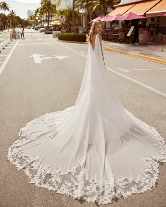 Een elegante trouwjurk van Rosa Clará’s Luna Novias collectie, model Camila. Deze jurk heeft een minimalistisch design met een aansluitende pasvorm, een subtiele split aan de voorkant, een lage ruglijn, en is afgewerkt met een afneembare cape met delicate kanten details.