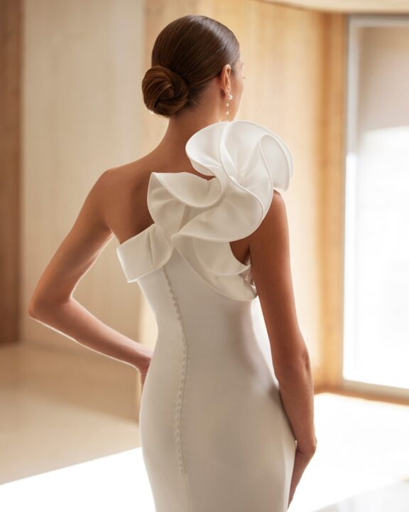 Elegant en modern design trouwjurk van Rosa Clará, model Abigail. Deze jurk heeft een opvallend asymmetrisch detail met een volumineuze ruches op één schouder. Gemaakt van soepel vallende crêpestof, met een aansluitende pasvorm die de vrouwelijke vormen accentueert. De achterkant is afgewerkt met een rij subtiele knoopjes en een sleep die de look compleet maakt.