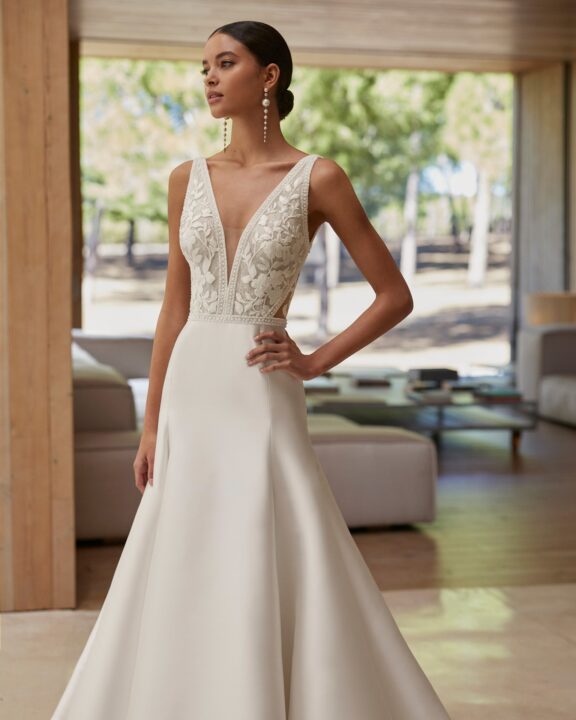 Elegante trouwjurk van Rosa Clará met een diep V-halslijn en kanten lijfje. De jurk heeft een volumineuze A-lijn rok van luxe mikado stof en een open V-rug met subtiele knoopsluiting. Perfect voor de moderne bruid die een tijdloze en verfijnde uitstraling zoekt.