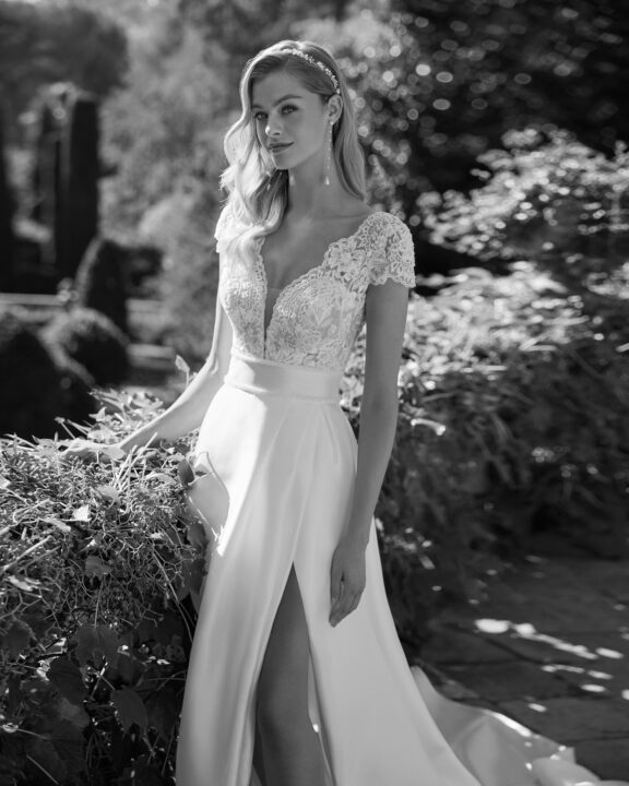 Elegante trouwjurk van Adriana Alier met kanten top, korte mouwen en een diepe V-hals. De soepelvallende satijnen rok heeft een hoge split en een lange sleep. Ideaal voor bruiden die een combinatie van romantiek en moderne elegantie zoeken.
